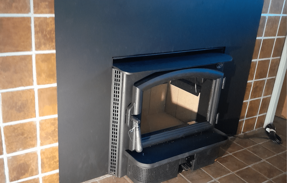 A black wood stove insert with its door open, set in a fireplace surrounded by brown and white tiles.